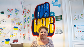 A photograph of a woman with short hair standing in front of a painted mural that reads, NO WAR in the colors of the Ukrainian flag.