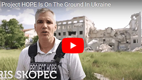 A screenshot from a video update from Project HOPE Vice President Chris Skopec when he recently vistied Ukraine. He is wearing a protective vest and polo shirt as he stands against the backdrop of the ruins of a destroyed building in a field of grass.