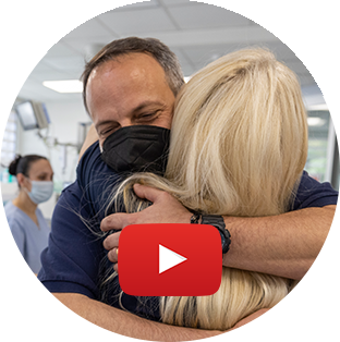 Project HOPE CEO Rabih Torbay smiles under his medical mask as he hugs a Ukrainian team member in a hospital. A large play button is shown on top of the photo.