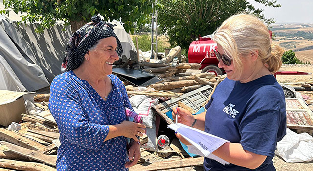 A photograph of an older woman in a patterned dress and head scarf smiles at a Project HOPE team member in sunglasses as they talk against the backdrop of fields, trees and a water access point.