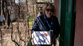 A photograph of a Project HOPE team member, wearing sunglasses and a jacket, as she carries a box containing humanitarian supplies into a brightly painted home in Ukraine.