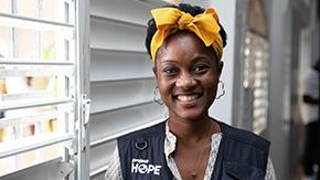 A photograph of a doctor in Hait wearing a Project HOPE vest, bow headband and earrings, as she smiles brightly against the backdrop of a hospital window covered with large blinds.