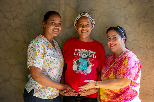 A photo of Yerika with the doctor and health worker supported and trained by Project HOPE. They all smile brightly at the camera as the doctor and health worker hold Yerika's pregnant belly.