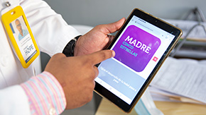 A photograph of a Project HOPE-supported health worker, wearing a nametag and lab coat, as they hold an ipad displaying text in Spanish reading, MADRE.