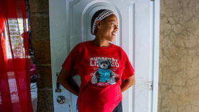 A photograph of Yerika as she stands against the door of her home in the Dominican Republic. She is wearing a t-shirt and hair covering as she smiles and looks to her right.