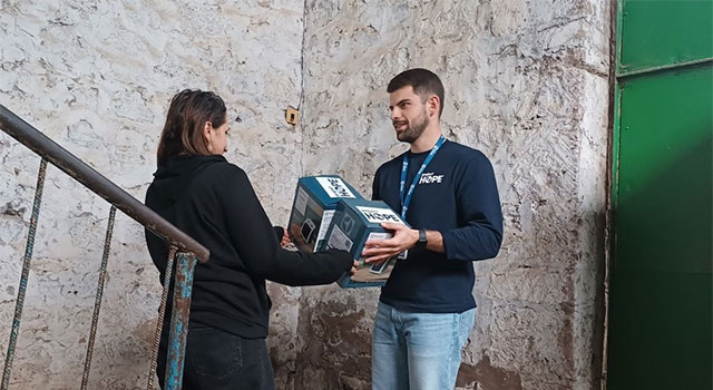 A photograph of Project HOPE team member in jeans and long sleeve shirt handing 2 small heaters to a woman standing on stairs against a textured white wall in Armenia.