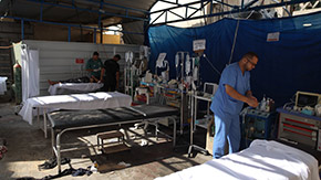 A photograph of a makeshift treatment room outside in Gaza, showing health workers preparing supplies next to gurneys on a dirt floor.