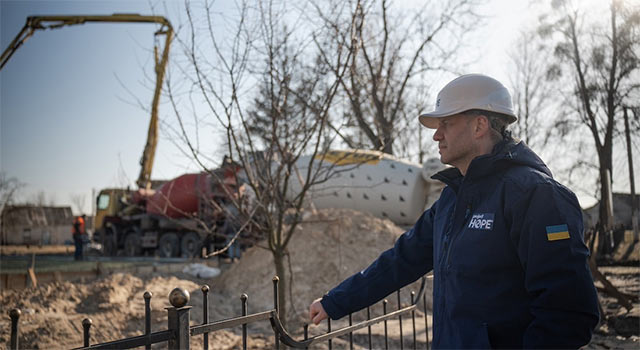 A photograph from Ukraine shows a man in a heavy jacket and construction helmet as he looks over a construction site, including a crane and cement truck against the backdrop of a few sparse trees.