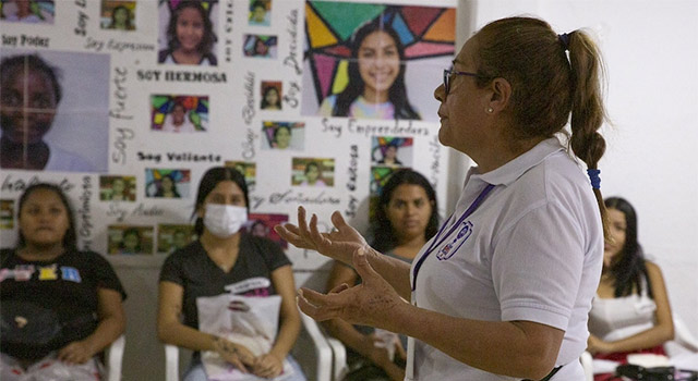 A photograph of Project HOPE partner in a polo shirt, glasses and ponytail in Colombia extends her hands out as she speaks to a group of young women sitting in plastic chairs as they listen.