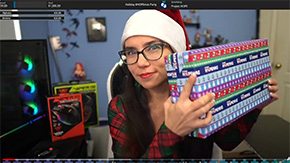 A screenshot of video showing a young woman in glasses and a plaid shirt as she holds up a present wrapped in Project HOPE-themed wrapping paper and wears a santa hat during a charity stream for Project HOPE.