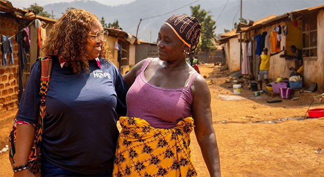 A photograph from Sierra Leone shows two women walking down a dirt path between rows of brick houses. One wears a Project HOPE shirt and glasses and the other, with her arm around her, wears a bandana and pink top.