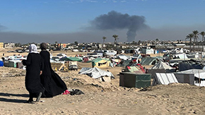 A photograph of two women wearing head scarves in Gaza walking over uneven sand against the backdrop of hundreds of tents of displaced people with a few trees. Smoke rises in the sky behind them.