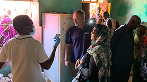 A photograph of Project HOPE's CEO and partners as they visit health facilities in Sudan. A doctor holds up a bubble pack of medicine in a green room where people wait outside to receive medications.