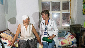 A photograph of a Project HOPE health worker from a mobile medical unit in Ukraine wears a stethoscope and carries a bag as she sits on a bed with an elderly woman who is wearing a bandana. They look at each other as they speak.