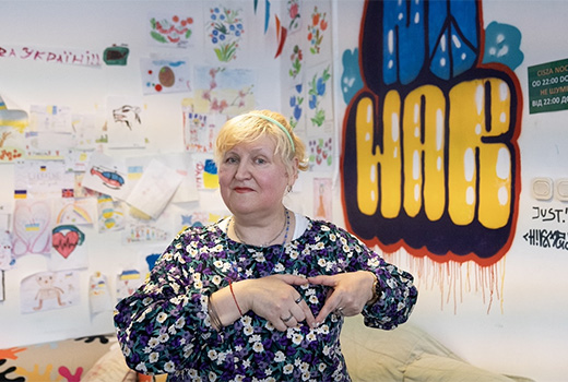 A photo of an older refugee from the Ukraine conflict at a Project-HOPE supported community center. She is wearing a floral long sleeve shirt, beaded necklaces and bracelets as she makes a heart symbol with her hands against her chest, against the background of children's artwork on the wall and a large graffiti-style mural reading, No War.