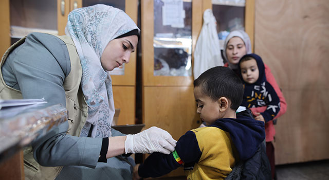 A photograph from a Project HOPE clinic in Gaza of a health care worker in a scarf, vest and gloves looking down intently as she measures the width of young boy's arm to determine if he's malnourished. The boy wears a hooded sweatshirt and also looks down at his arm as his family waits in the background.