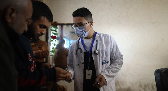 A photograph of a Project HOPE-supported health worker in Gaza as he hands out a blister pack to a man holding a small child at a short-term medical clinic for displaced families. He wears a doctors coat, medical mask, stethscope and glasses.