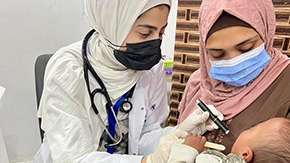 A close-in photograph from Project HOPE's primary health clinic in Gaza shows two women in headscarves and medical masks, one is holding a baby, and the other is a health care worker using a light to look into the baby's mouth.