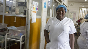 A photograph of a health worker in Sierra Leone smiles brightly as and wears scrubs and a medical cap as she walks down a hallway of the Special Care Baby Unit at Ola During Children's Hospital.