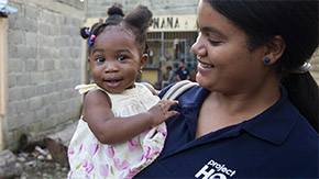 A photograph of a Project HOPE health worker in a polo is shown in the Dominican Republic as she holds a smiling baby in a dress with small beads in her hair. They stand outside against the backdrop of a brick wall.