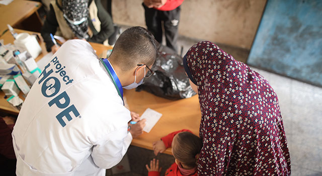 A photograph from a Project HOPE clinic in Gaza of a health care worker in a lab coat and mask as he writes on a pad of paper for a mother and child who he is helping in a clinic for displaced people in Gaza.