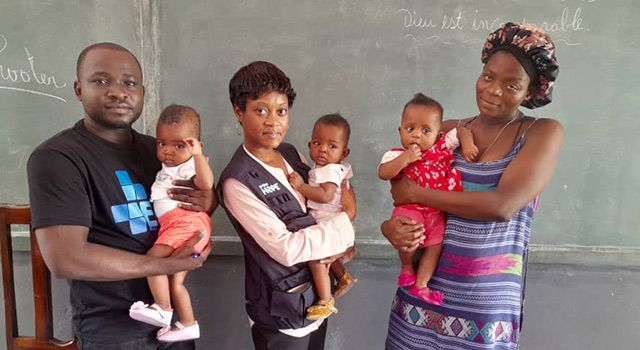 A photograph of a Project HOPE mobile clinic in Haiti where two health workers and a mother each hold a young triplet as they stand in front of a blackboard.