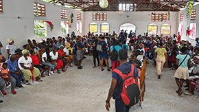 A photograph of a large group of people in Haiti waiting for health care services in a large white room with an open wood beam roof.