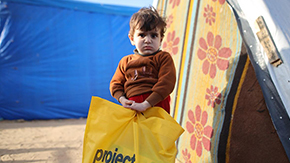 A photograph from a Project HOPE distribution to displaced people in Gaza, showing a young boy with rumpled hair holding a yellow Project HOPE bag with aid supplies as they look at the camera.