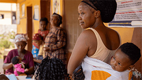 A photograph of a woman with her baby on her back as she looks to the left with a slight smile on her face. The background is at a Project HOPE-supported clinic in Sierra Leone where many other women and their children are waiting.