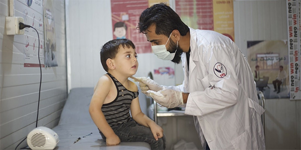 Photograph from Syria shows a doctor performing an exam on a young child wearing a tank top and jeans as he sits on an exam table in a clinical setting.