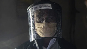 A health worker in India wears a steamed up face shield and medical mask and a light blue collared shirt as he looks to a colleague to consult in a dark room.