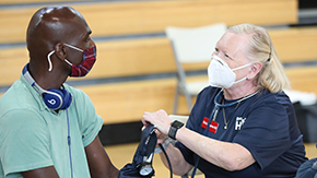 A photograph of Project HOPE volunteer in a navy blue shirt, black watch and white mask as she's about to take the blood pressure of a Hurricane Ida survivor, also in a mask and a green shirt with blue headphones.