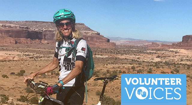 A person withe a green helmet and black and white biking outfit balances on her black and white bike as she holds the handlebars and smiles while wearing dark sunglasses. She has blonde hair and is surrounded by a mountainous desert landscape