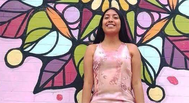 A woman in Mexico wears a pink printed tank top and her long dark hair is down, pushed back behind her shoulders. She smiles as she stands in front of a brick wall painted pink and purple with a brightly-colored geometric and floral mural.