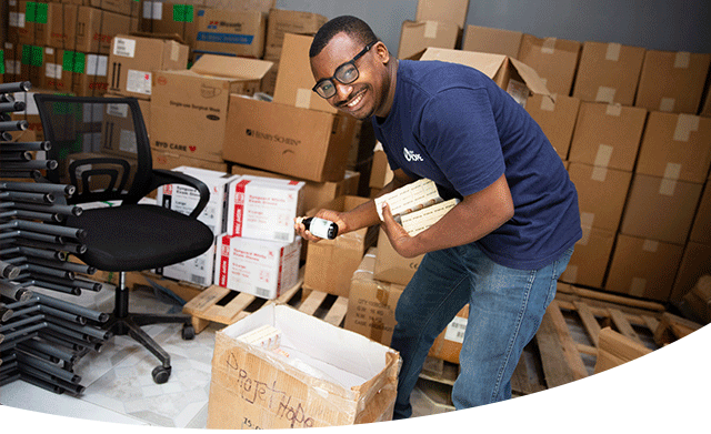 A health worker is in Haiti, in a room filled with cardboard boxes full of medical supplies. He smiles brightly as he leans over an open box and holds several small packages in his arm and a small medicine bottle in his hand. He is wearing jeans, a Project HOPE shirt and glasses. 