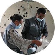 A photograph of two health workers wearing light blue face masks and Project HOPE vests clean up a clinic. They stand in front of a wall full of holes from violence in the region.