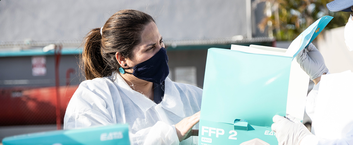 A health care worker with her hair in a pony tail, wears a black face mask and protective clothing as she squints to examine a box of medical supplies.