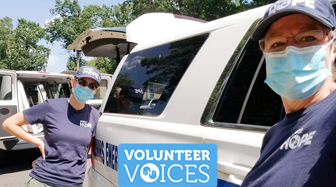 Two women in Project HOPE gear and masks lean against against an emergency vehicle and make eye contact with the camera. 