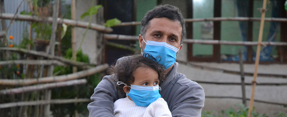 A father and child, both wearing medical blue face masks, are seated on straw bales in front of a rustic wooden fence and a garden. The father, with a solemn look at the camera, has his young child in his arms as she touches his hand and looks left, out to the world beyond.