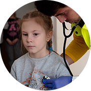 A photograph of young Ukranian refugee wearing a gray sweatshirt getting her breathing checked with a stethoscope by a health worker in a yellow shirt and vest. 