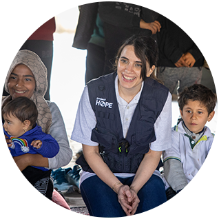 A photograph of a Project HOPE team member shows her smiling and sitting in a safe space for children after the traumatic earthquakes in Turkiye and Syria. On one side of her is a laughing young girl holding a baby, and on the other side sits a young boy wearing a polo and long sleeves who looks serious as he watches the activities.