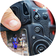 A photograph of a camera held by a person's hand, showing up close buttons and the screen previews a photo in progress of a family sitting on brick stairs. 