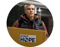 A man with salt and pepper hair wears a winter vest and jacket as he carries a box of supplies labeled with the Project HOPE logo into a hospital in Ukraine.