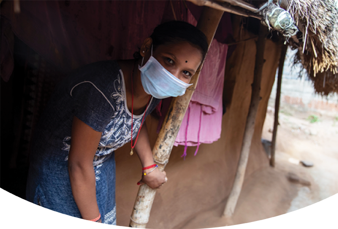 A young person, wearing a white-embroidered blue dress, red corded necklace and bracelet, smiles under her medical mask as she walks out of a small home with a straw roof. She is in a village in India where Project HOPE supplies helped the community reduce their new infection rate to zero.