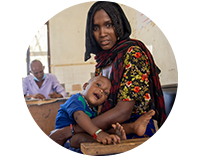 A mother wearing a head scarf looks at the camera while holding her young child in a tshirt and shorts who smiles playfully as a doctor works in the background.