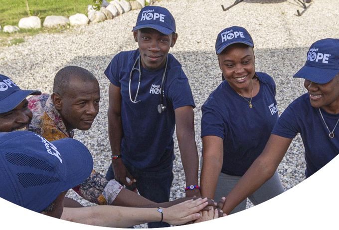 A group of medical volunteers in Project HOPE navy blue stand in a circle with hands in. Everyone is smiling as they get ready to help earthquake survivors in Haiti.