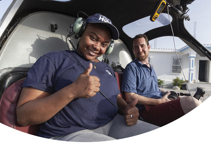 Nurse Lyronne Sam, left, and Tom Cotter, right, are part of Project HOPE's emergency response team in Haiti. They both wear navy shirts and smile at the camera as they sit in a helicopter in Haiti. Nurse Sam holds up her hand with a thumb's up.