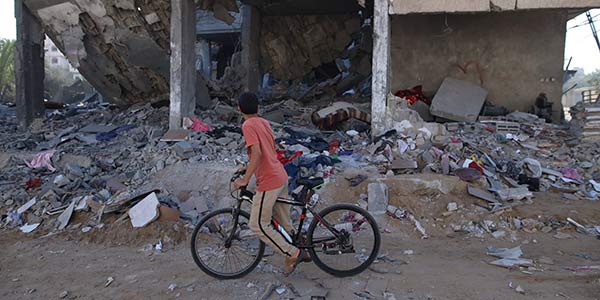 A photograph from Gaza shows a young boy riding his bike by and looking at a completely destroyed building filled with debris and walls no longer standing.