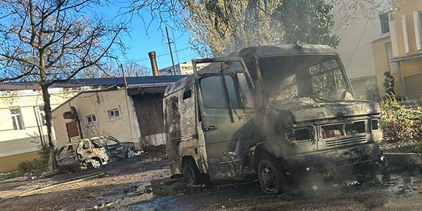 Photograph showing artillery damage to a small van and another sedan outside a hospital in Ukraine. Steam rises from the vehicles.