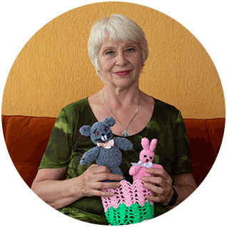 Round photo of a Ukrainian refugee named Galina as she holds two knit toy animals, a bunny and mouse, she made at the community center. She has short hair, a kind smile and is sitting on a couch.